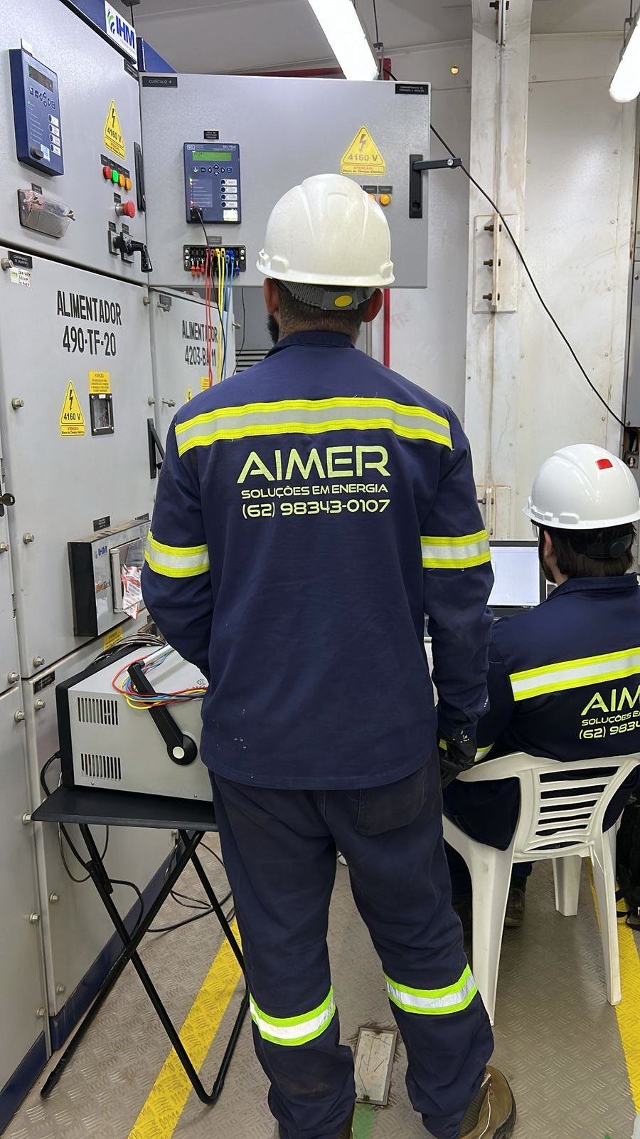 Técnico da Aimer, de uniforme e capacete, trabalhando diante de painéis elétricos
